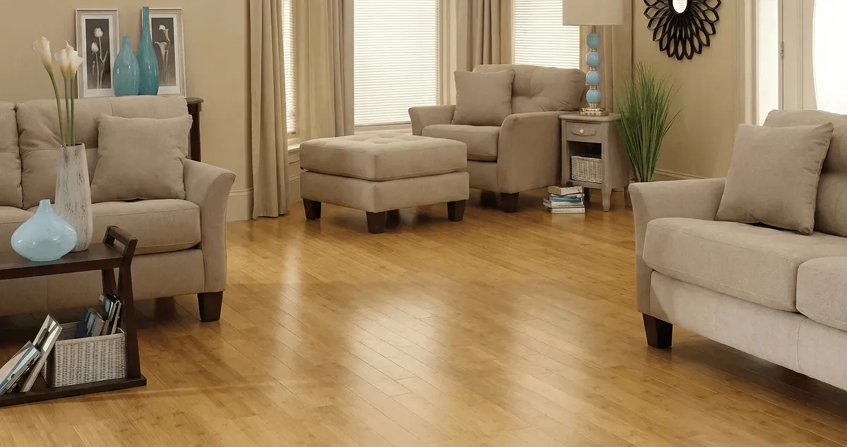 A living room with professionally installed bamboo flooring, beige sofas, a matching ottoman, a side table with a blue lamp, vases, framed pictures, and large windows with blinds and curtains in Fort Worth, TX